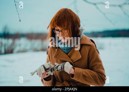 Porträt einer jungen, rothaarigen Asiatin, die an einem frostigen Wintertag im Park Wollhandschuhe auszieht. Nach Norden. Stockfoto