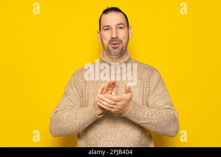 bärtiger lateinamerikanischer Mann mit beigefarbenem Rollkragenpullover, der für gute Nachrichten klatscht, isoliert auf gelbem Hintergrund Stockfoto