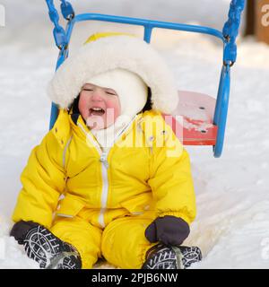 Das Kleinkind fiel von der Schaukel in den Schnee auf dem Winterspielplatz. Ein Junge im gelben Schneeanzug, der im Kindergarten weint. Ein Kind im Alter von einem Jahr, acht Monate Stockfoto