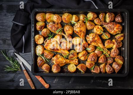 Gebackene heiße Hähnchenschenkel mit würzigen Kartoffeln in Backblech auf schwarzem Hintergrund Stockfoto