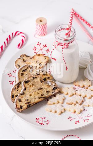 Flasche Milch mit traditionellem Weihnachtsstollen, kleinen Keksen und Zuckerrohr auf weißem Hintergrund. Deutsches Weihnachtsbrot Stockfoto