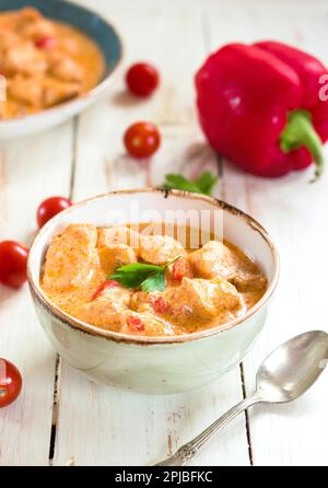 Köstlicher Hühnereintopf mit Paprika in einer Schüssel auf einem weißen Holztisch. Mit frischen Kirschtomaten, roter Paprika und Petersilie. Traditionelles ungarisch Stockfoto