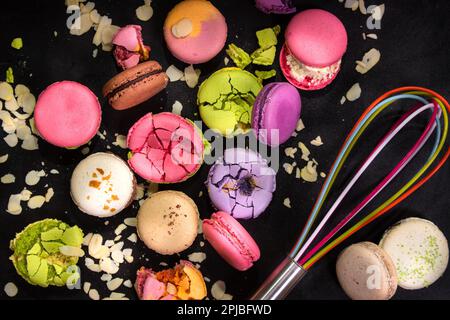 Bunte französische Makronen und Mandelflocken mit Schneebesen auf schwarzem Hintergrund. Nahaufnahme. Draufsicht. Konzept der Backmakronen Stockfoto