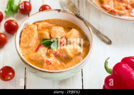 Köstlicher Hühnereintopf mit Paprika in einer Schüssel auf einem weißen Holztisch. Mit frischen Kirschtomaten, roter Paprika und Petersilie. Traditionelles ungarisch Stockfoto