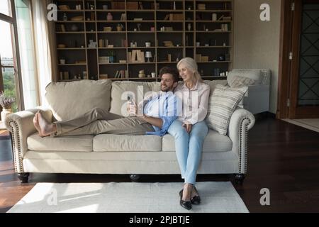 Ein glücklicher junger Erwachsener ruht sich auf dem Sofa neben der älteren Mutter aus Stockfoto