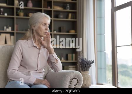 Deprimierte, einsame, ältere, grauhaarige Frau, die auf der Heimcouch sitzt Stockfoto