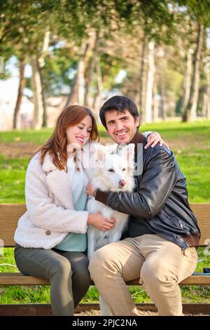 Ein junges weißes Paar streichelt seinen Hund auf einer Bank in einem Park. Stockfoto