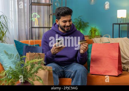 Glücklicher, einkaufsamer, indischer Konsumenten, der mit Einkaufstaschen in einem modernen Apartment sitzt und online bestellt, mit Kreditkarte bezahlt. Rabattangebot im Internet. Junger hindu-Typ auf dem Sofa Stockfoto