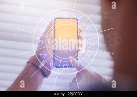 Mann mit Smartphone liest täglich Horoskop drinnen, Nahaufnahme Stockfoto