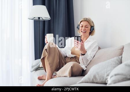 Eine Frau mittleren Alters in eleganter Kleidung ist zu Hause und benutzt ihr Telefon Stockfoto