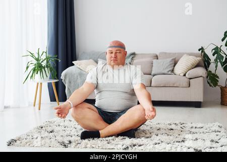 Yoga-Übungen. Der lustige, übergewichtige Mann in legerer Kleidung ist zu Hause Stockfoto