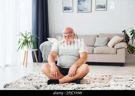 Yoga-Übungen. Der lustige, übergewichtige Mann in legerer Kleidung ist zu Hause Stockfoto