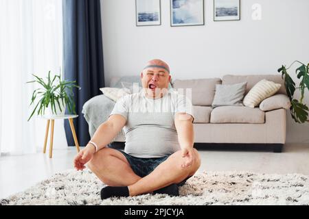 Yoga-Übungen. Der lustige, übergewichtige Mann in legerer Kleidung ist zu Hause Stockfoto