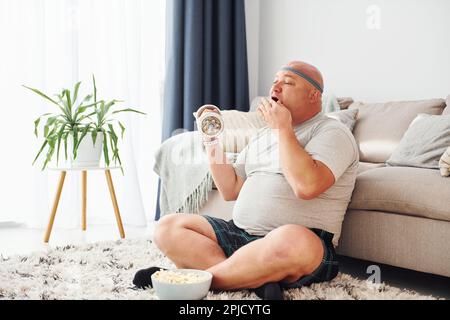 Er isst und trainiert. Der lustige, übergewichtige Mann in legerer Kleidung ist zu Hause Stockfoto