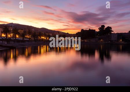 Der Blick auf die Stadt über das stille Wasser (Meer) in Coullioure, Frankreich nach dem Sonnenuntergang. Pink- und Violetttöne, der Himmel reflektiert sich im Wasser. Stockfoto