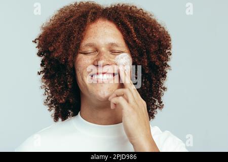 Ein Mann mit einem geschwungenen Afro, der seine Haut mit einer feuchtigkeitsspendenden Gesichtscreme pflegt. Ein hübscher junger Mann, der seine Hautpflege mit einem großen Lächeln praktiziert. Stockfoto