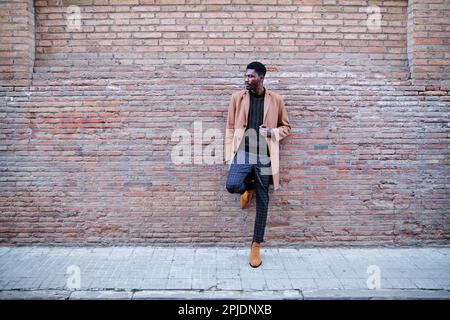 Eleganter afroamerikanischer Mann, der sich draußen auf der Straße gegen eine Backsteinmauer lehnt. Stockfoto