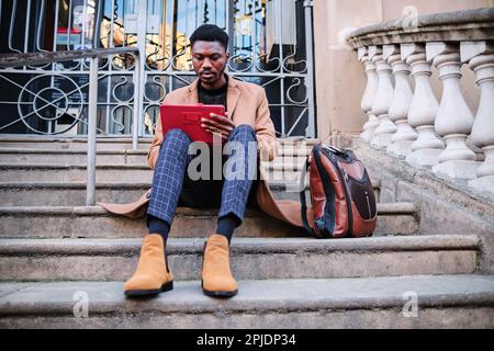 Eleganter schwarzer Mann, der ein digitales Tablet benutzt, während er draußen auf Treppen sitzt. Technologiekonzept. Stockfoto