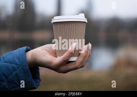 Trinken Sie Kaffee in der Natur aus einem ökologischen Pappbecher. Hände erwärmen, während Sie eine Tasse mit einem heißen Getränk halten, gehen Sie, um sich zu beruhigen. Stockfoto