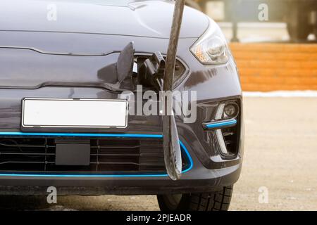 Elektroauto an Ladestation angeschlossen, um Batterie aufzuladen, Detail, Vorderansicht, zusätzliches Sonnenlicht. Keine Personen, Logos usw. entfernt. Stockfoto