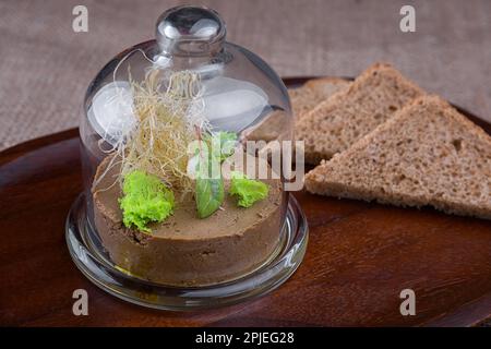 Reichhaltige und cremige Leberpastete mit Kräutern und getoastetem Brot unter einer Glaskuppel Stockfoto