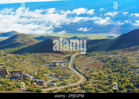 Besucherbereich inmitten des Tals von Mauna Kea mit kurvenreichen Straßen, vulkanischen Hügeln und Wolken aus der Ferne. Stockfoto