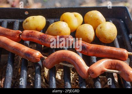 Köstliche Würstchen und Kartoffeln, die über einem offenen Feuer geröstet werden, mit einem Grillgitter, um sie beim Kochen sicher zu halten. Essenz der Outdoor-Küche. Stockfoto