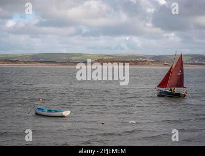 Segelboot mit roten Segeln neben einem blau-weißen Ruderboot, das im Wasser verankert ist, abgebildet vom Appledore Harbour in North Devon im Oktober Stockfoto