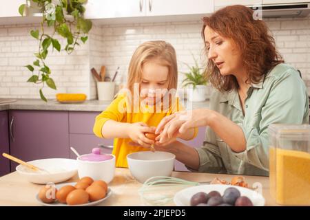 Großmutter und Enkelin bereiten Kuchenteig zu, verbringen Zeit miteinander, Familienbeziehungen, Liebe. Geben Sie ein Ei zum Teig Stockfoto