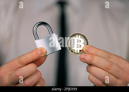 Ein Geschäftsmann hält ein silbernes Vorhängeschloss und eine physische Version von Bitcoins in seinen Händen. Verbot von Kryptowährungen, Vorschriften, Beschränkungen oder Sicherheit Stockfoto