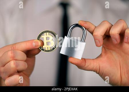 Ein Geschäftsmann hält ein silbernes Vorhängeschloss und eine physische Version von Bitcoins in seinen Händen. Verbot von Kryptowährungen, Vorschriften, Beschränkungen oder Sicherheit Stockfoto