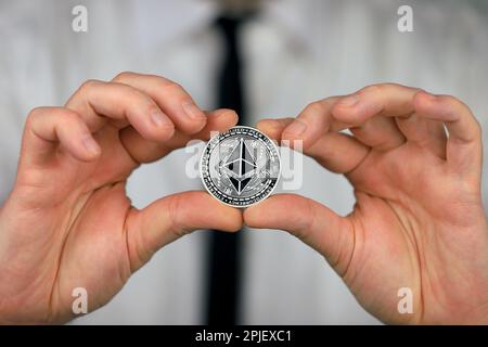 Ein Geschäftsmann mit weißem Hemd und schwarzer Krawatte hält eine physische Version von ethereum in seinen Händen. Stockfoto