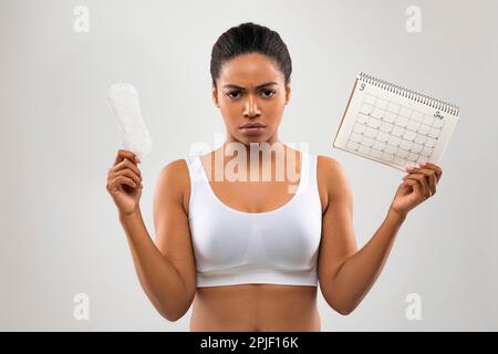 Wütende junge schwarze Frau zeigt Periodenkalender und hygienische Serviette Stockfoto