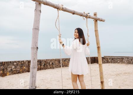 Glückliche attraktive junge asiatische Frau in weißem Kleid, die im Sommer eine Holzschaukel am Strand spielt Stockfoto