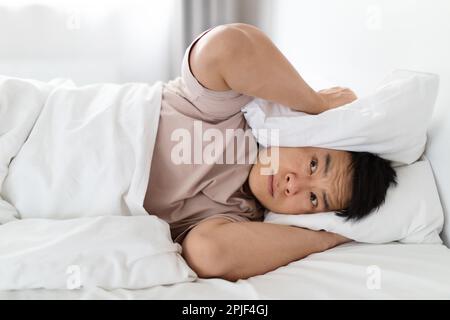 Schlafloser Mann lag im Bett und bedeckte die Ohren mit einem Kissen Stockfoto
