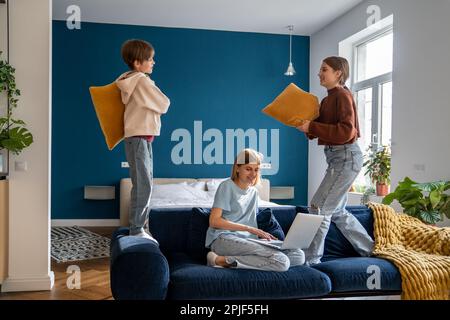 Mutter, die an einem Laptop arbeitet, während ihre aktiven, lauten Kinder mit Kissen spielen und Spaß haben Stockfoto