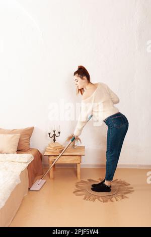 Eine schöne junge Frau putzt das Haus und hört Musik. Beigefarbenes Interieur, komisches Mädchen. Eine Frau wäscht den Boden mit einem Mopp. Stockfoto