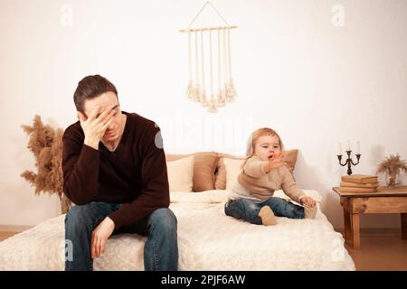 Dad kommt mit dem Kind nicht klar, ein zweijähriges Mädchen liegt auf dem Bett und schreit hysterisch. Vater müde unglücklich. Ein Mann ist allein zu Hause Stockfoto