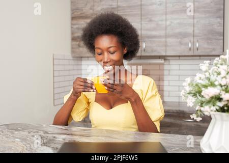 Eine wunderschöne afroamerikanische Frau trinkt Tee in der Küche. Genießen Sie ein Getränk, heißen Kaffee am Morgen, Frühstück. Energie- und Lebenserwartung für den ganzen da Stockfoto