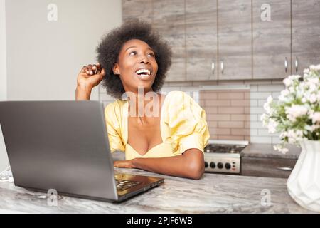 Porträt einer wunderschönen afroamerikanischen Freiberuflerin, Tisch in der Küche. Arbeiten Sie mit dem Laptop, drinnen. Lacht Stockfoto