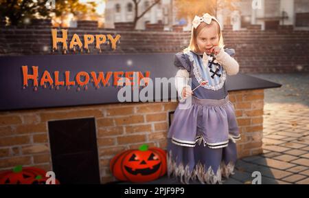 Kleines hübsches Mädchen, Kunstmagie Postkarte, künstlerisch. Banner mit Text Happy Halloween, glühende Kürbisse. Kind in Hexenkostüm, isst süße Kand Stockfoto