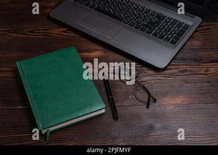 Grünes Tagebuch auf dem Tisch. Tagebuch und Laptop. Notizblock, Laptop und Brille. Führen Sie einen Tagebucheintrag. Draufsicht in einem Winkel. Stockfoto