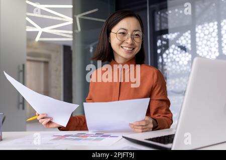 Junger erfolgreicher asiatischer Finanzier hinter Papierarbeit im Büro, Buchhalterin der Geschäftsfrau, die Vertragsdokumente und Berichte lächelnd und glücklich prüft, zufrieden mit den Ergebnissen, die mit dem Laptop erzielt wurden. Stockfoto