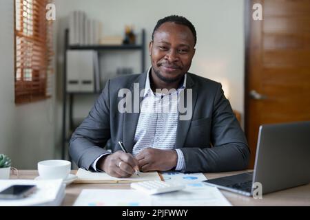 Ein Mann im mittleren Alter, der ein amerikanischer Afrikaner ist, der einen Computer-Laptop verwendet, um an Finanzdokumenten, Steuern, Wechselgeld, Buchhaltung und Finanzberatern zu arbeiten Stockfoto