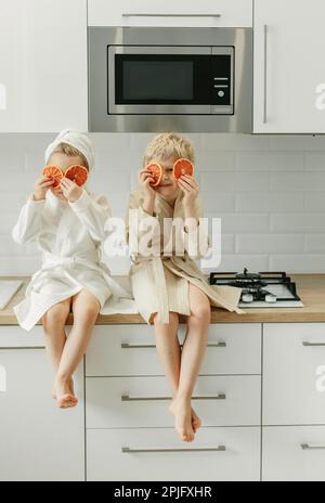 Ein Mädchen und ein Junge in Bademänteln sitzen in der Küche und schließen ihre Augen mit kandierten Orangen. Stockfoto