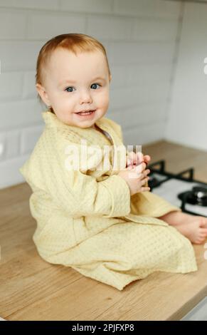 Ein Mädchen im gelben Bademantel sitzt auf dem Küchentisch und isst Marmelade. Stockfoto