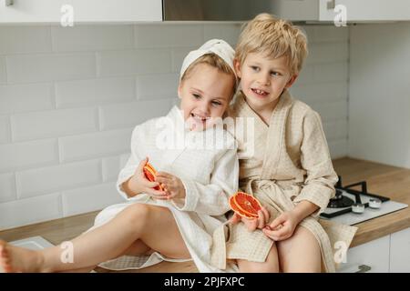 Ein Mädchen und ein Junge in Bademänteln sitzen in der Küche und schließen ihre Augen mit kandierten Orangen. Stockfoto