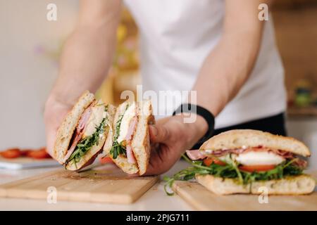 Ein hübscher junger Mann schneidet italienisches Sandwich und zeigt, was drinnen ist. Köstliches Mittagessen zu Hause Stockfoto