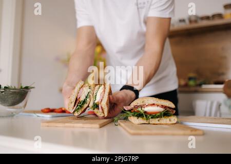 Ein hübscher junger Mann schneidet italienisches Sandwich und zeigt, was drinnen ist. Köstliches Mittagessen zu Hause Stockfoto