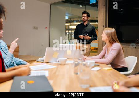Selbstbewusster männlicher Sprecher hält eine Präsentation für verschiedene Geschäftsleute im Konferenzraum und spricht über einen neuen Geschäftsplan. Startup-Team sitzt bei Stockfoto